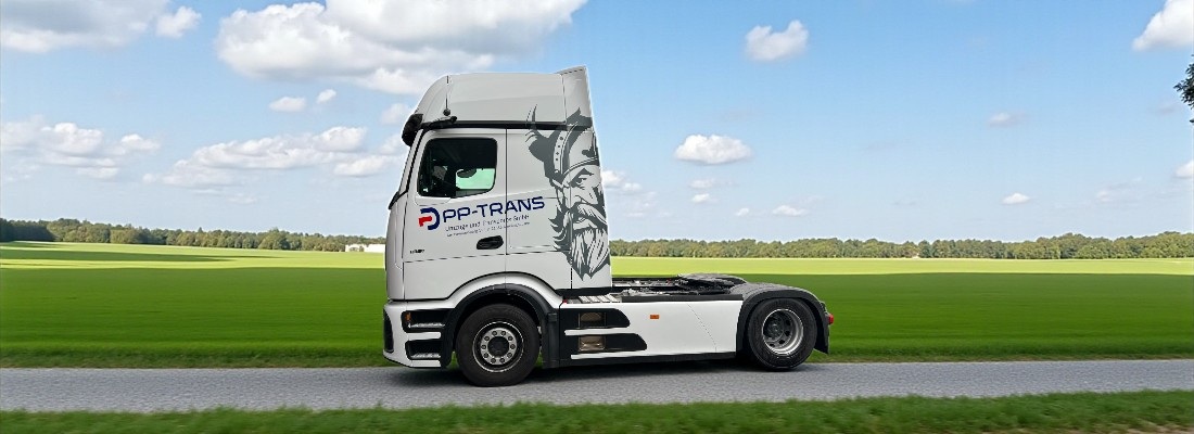 Ein weißer Sattelzug-Truck ohne Auflieger fährt auf einer schmalen Straße durch eine grüne, ländliche Landschaft. Auf der Fahrertür ist ein Logo mit der Aufschrift „PP-TRANS“ sowie ein stilisiertes Bild eines Wikingerkopfs zu sehen. Im Hintergrund erstrecken sich weite grüne Felder und ein Waldstreifen unter einem blauen Himmel mit einigen weißen Wolken.