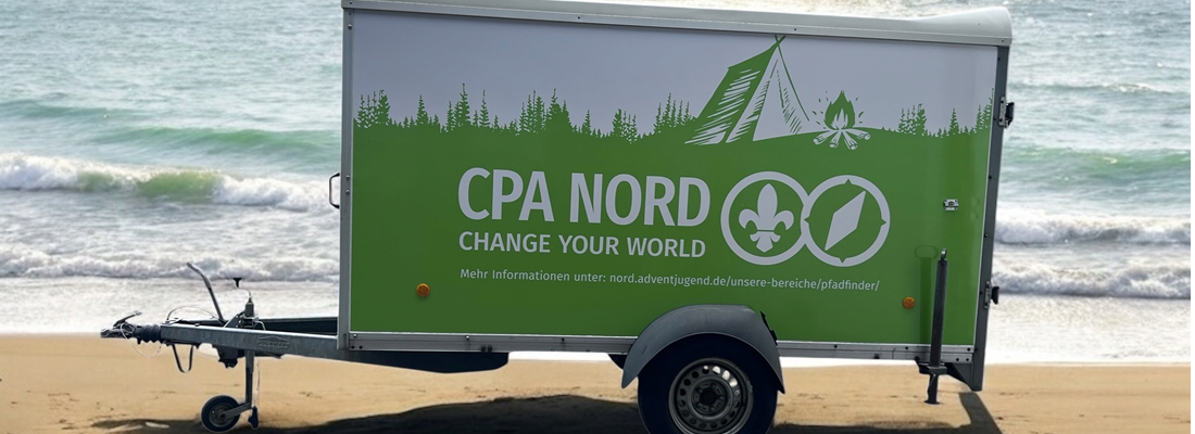 Ein grün-weißer Anhänger mit dem Text „CPA NORD CHANGE YOUR WORLD“ und Campingsymbolen steht an einem Sandstrand in der Nähe des Ozeans.