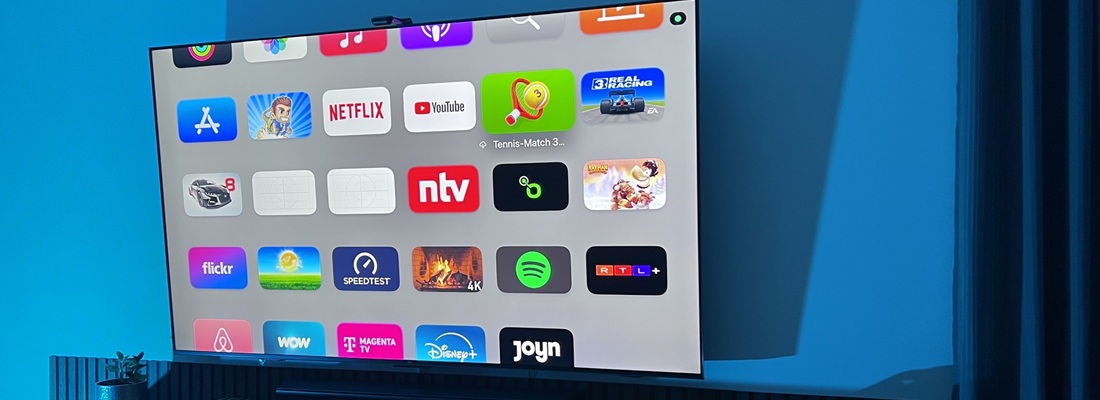 Fernseher mit Apple-TV-Oberfläche zeigt verschiedene Streaming- und App-Icons wie Netflix, YouTube, Disney+, Spotify, RTL+ und mehr. Der Bildschirm ist von einem kühlen, blauen Hintergrundlicht umgeben, das an eine LED-Umgebungsbeleuchtung erinnert. Der TV hängt an der Wand über einem Lowboard.