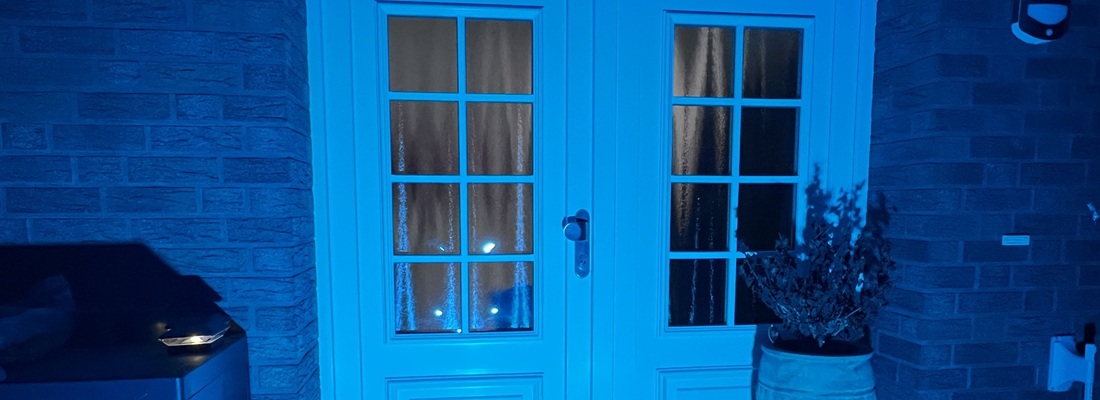 Zweiflügelige weisse Terrassentür mit Glasfenstern in Gitteraufteilung, beleuchtet von blauem Licht. Links neben der Tür steht ein Grill oder Metallgerät, rechts ein grosser Blumentopf mit einer vertrockneten Pflanze. Die Tür ist in eine Fassade aus dunklen Ziegelsteinen eingebaut.