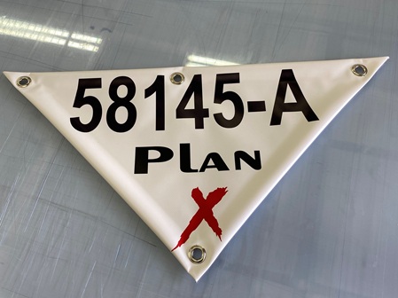 Ein rechteckiges, weißes Banner mit einem roten „X“ und der Aufschrift „58145-A PLAN“ in großen, schwarzen Buchstaben. Das Banner hat an den oberen Ecken Metallösen, die zur Befestigung dienen. Die Oberfläche des Banners ist glatt, und im Hintergrund sind Lichtreflexionen sichtbar, was auf eine glatte oder glänzende Oberfläche hinweist.