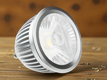 Nahaufnahme einer LED-Spotlampe mit silbernem Kühlrippengehäuse und transparenter Abdeckung, die einen Reflektor und das LED-Leuchtmittel im Innern zeigt. Die Lampe liegt leicht geneigt auf einer Holzoberfläche, im Hintergrund eine verschwommene, braune Wand mit dunklen Punkten.