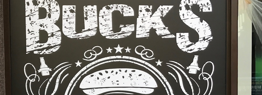 Schaufensterbeschriftung Hamburg: auffälliger Schriftzug BUCKS mit Burger-Logo, Sternen und Bierflaschen als Folienplott für Gastronomie
