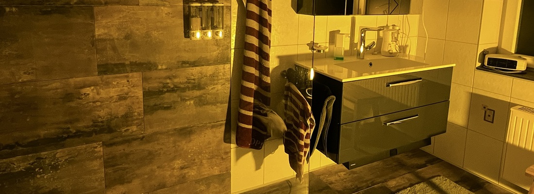 Badezimmer mit warmgelbem Licht, vermutlich durch eine Farbfolie verändert. Links eine Dusche mit Wandfliesen in Betonoptik, in der Mitte ein Spiegel mit Handtüchern und Spendern, rechts ein modernes Waschbecken mit Unterschrank. Das gelbliche Licht verleiht dem Raum eine warme Atmosphäre.