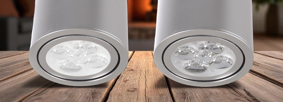 Zwei zylindrische LED-Deckenstrahler mit silbernem Gehäuse, nebeneinander auf einem Holzboden stehend und von unten fotografiert. In jedem Strahler sind vier LED-Linsen sichtbar. Im Hintergrund ist unscharf eine warme Innenraumszene zu erkennen.