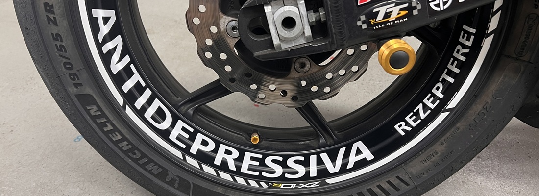 Nahaufnahme eines Motorrad-Hinterrads mit auffälliger weisser Beschriftung auf der schwarzen Felge: „ANTIDEPRESSIVA REZEPTFREI“. Auf dem Reifen ist der Markenname „MICHELIN“ sowie die Reifengrösse zu erkennen. Sichtbar sind zudem Bremsscheibe, Felgenventil und Teile der Radaufhängung.