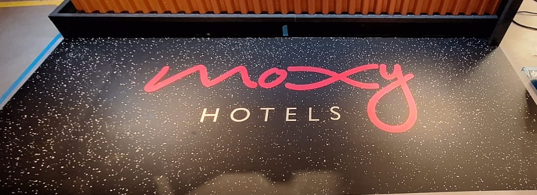 Eingangsbereich mit schwarzer Bodenbeschriftung: Moxy Hotels Logo in Pink auf Folienboden mit weißen Sprenkeln