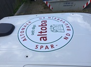 Logo auf dem Dach eines weissen Fahrzeugs mit der Aufschrift „altoba – seit 1892“. Der Schriftzug ist kreisförmig angeordnet und lautet vollständig: „Altonaer Spar- und Bauverein eG“. In der Mitte des Logos sind stilisierte Figuren eines Paares in historischer Kleidung abgebildet. Im Hintergrund ist ein Abfallcontainer mit einer Webadresse und Telefonnummer zu sehen.