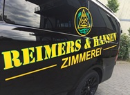 Schwarzer Firmenwagen mit der auffälligen gelb-grünen Beschriftung "REIMERS & HANSEN ZIMMEREI" auf der Seite. Daneben befindet sich ein rundes Zimmerei-Logo mit Winkelmass, Eichenblättern und Wellen. Das Fahrzeug steht auf einem gepflasterten Parkplatz vor einem Industriegebäude.