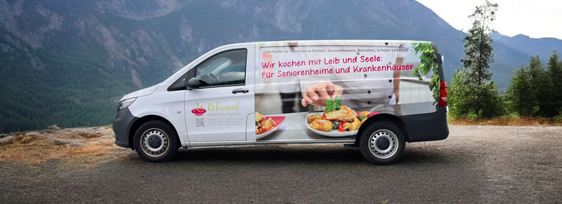Lieferwagen mit grosser Seitenbeklebung, auf der ein Koch beim Anrichten von Speisen zu sehen ist. Der Text auf dem Fahrzeug lautet: „Wir kochen mit Leib und Seele für Seniorenheime und Krankenhäuser“. Links befindet sich das Logo „Rebional“ mit zusätzlichen Informationen über die Zielgruppen wie Kitas, Schulen und Kliniken. Der Wagen steht auf einer Bergstrasse mit Blick auf bewaldete Berge im Hintergrund.