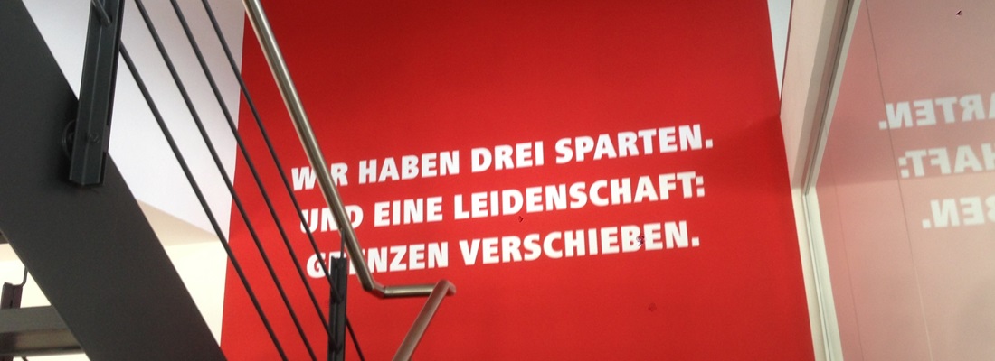Moderne Wandgestaltung im Treppenhaus mit roter Folienbeklebung und weißem Schriftzug „Wir haben drei Sparten. Und eine Leidenschaft: Grenzen verschieben.“ – umgesetzt von Folien Fischer Hamburg Bergedorf für Firmenräume in Reinbek, Glinde, Geesthacht, Schwarzenbek, Lauenburg und Wentorf.