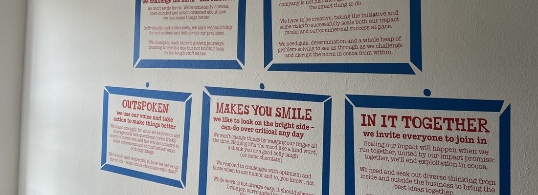 Sechs farblich gerahmte Plakate mit englischen Texten hängen an einer weissen Wand. Die Überschriften in roter Schrift lauten: „RAISE THE BAR“, „ENTREPRENEURIAL“, „OUTSPOKEN“, „MAKES YOU SMILE“ und „IN IT TOGETHER“. Jeder Titel ist mit einem erklärenden Text darunter versehen. Die Plakate sind mit blauem Klebeband befestigt und scheinen Unternehmenswerte oder Leitprinzipien darzustellen.