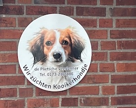 Rundes Schild an einer Backsteinwand mit dem Bild eines Kooikerhondje-Hundes in der Mitte. Darauf steht der Text: „Wir züchten Kooikerhondje“, sowie „de Plietsche Veehanner“ und eine Telefonnummer. Das Schild dient der Werbung für eine Hundezucht.