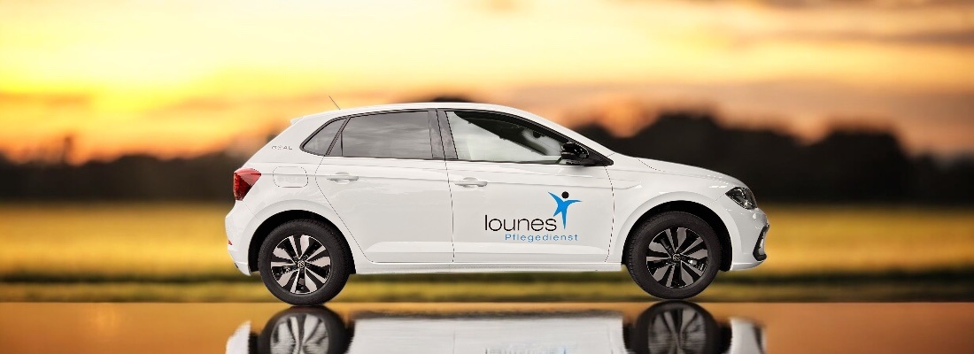 Weisser Kleinwagen mit der Aufschrift „lounes Pflegedienst“ und einem blauen stilisierten Menschen-Logo auf der Fahrertür, vor einem unscharfen Hintergrund mit Wiese, Bäumen und Sonnenuntergang. Das Auto spiegelt sich auf der glänzenden Oberfläche, auf der es steht.